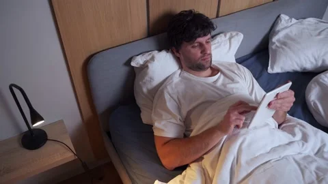 Young handsome man uses a tablet lying on the bed at night. A man suffering from Vídeos de archivo 233282424