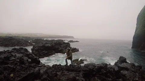 Young handsome man walking through the rocks on the shore of the sea in overcast Stock Footage