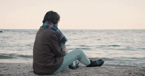 Young handsome man in warm coat sits by sea and looks at waves. Romantic Stock Footage 143814633
