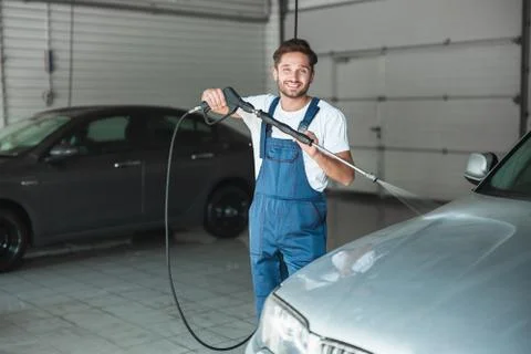 Young handsome man wearing uniform washing car at car washing station using high Stock Photos