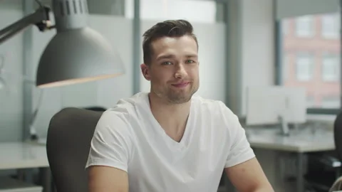 A young handsome man in a white T-shirt sits at an office desk and winks, slow Stock Footage 150333331