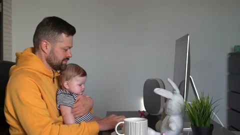 Young handsome man working on a desktop computer at home with a baby in his arms Stock Footage 151036132