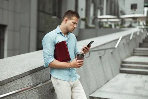 Young handsome office worker checking his smarphone holding laptop and cup of Stock Photos