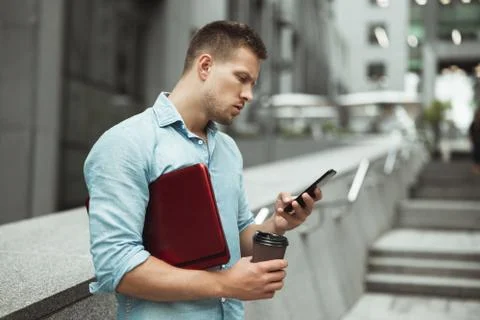 Young handsome office worker checking his smarphone holding laptop and cup of Stock Photos
