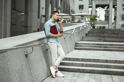 Young handsome office worker talking on the phone holding laptop standing on the Stock Photos