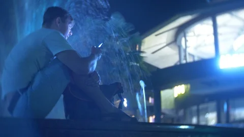 A young handsome sad guy sits in the evening next to a fountain with colored Видео 93206191