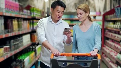 Young happy Asian couple using smartphone in supermarket with shopping cart choo Stock Footage 199846151