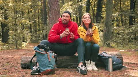 Young happy couple sitting on forest glade on log talking while drinking hot Stock Footage 147070923