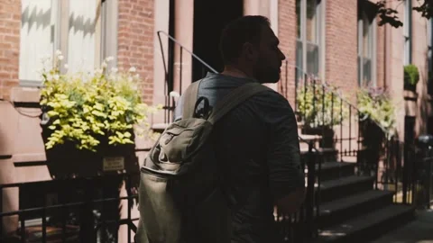 Young happy man with backpack walking along sunny summer Brooklyn street in New Vidéo 93948974