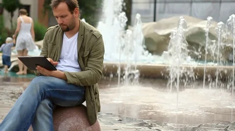 Young happy man with tablet computer sitting by the fountain, slow motion HD Stock Footage 8524029