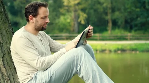Young happy man with tablet computer by the tree in park HD Stock Footage 8574531