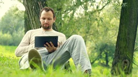 Young happy man with tablet computer by the tree in park, dolly shot Stock Footage 8581996