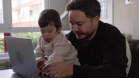 A young happy man trying to work on laptop holding his son at home. Remote work Stock Footage 245424393