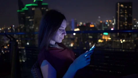 Young, happy woman texting on smartphone sitting on terrace in bar at night Stock Footage