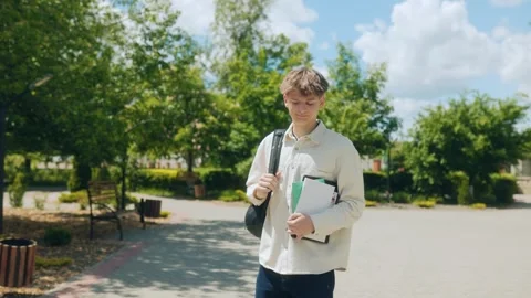 Young high school boy strolling in schoolyard holding study materials Stock Footage 310882530