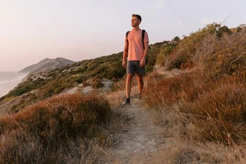 Young hiker man with backpack walking on the coast with warm sun light. Stock Photos