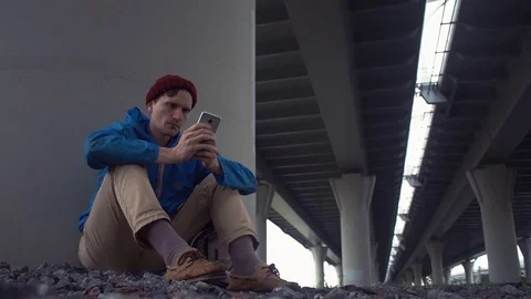 Young hiker using smartphone sitting on the ground under road bridge Stock Footage 80285868