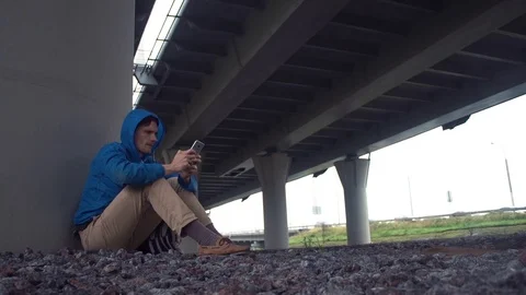 Young hiker using smartphone sitting on the ground under road bridge Stock Footage 80286127