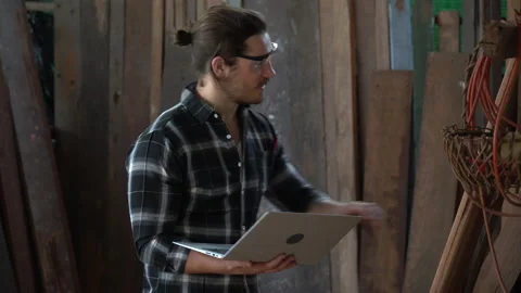 Young hipster carpenter man holding computer laptop looking and checking wood Stock Footage 149985174
