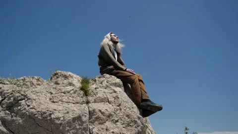 A young hipster guy with long blond hair sits on the edge of a cliff Video stock 147908031