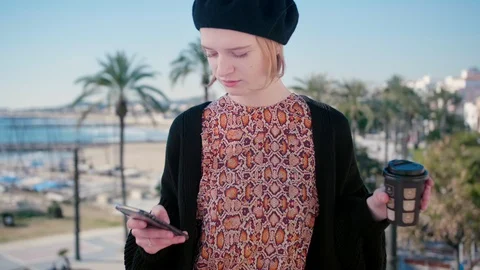 Young hipster lady with coffee using phone on holidays sunny sea town background Stock Footage 105607615