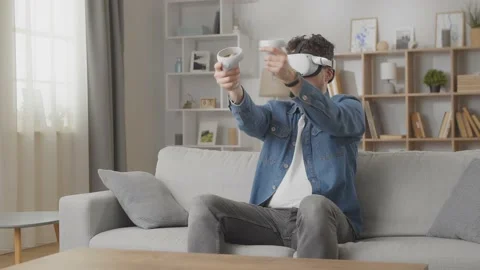 Young hipster man using his VR headset for playing games on sofa at home in the Stock Footage 169320743