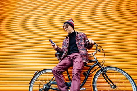 Young hipster man using mobile phone while sitting on cruiser bicycle near Stock Photos