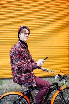 Young hipster man using mobile phone while sitting on city bicycle near orange Stock-Fotos