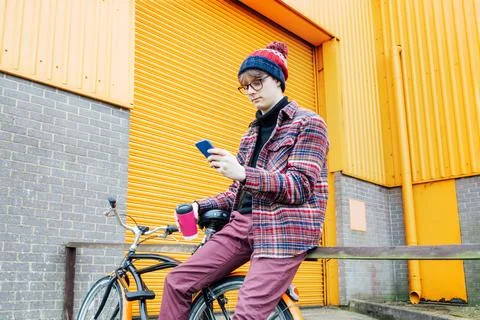 Young hipster man using mobile phone while sitting on cruiser bicycle near Stock Photos