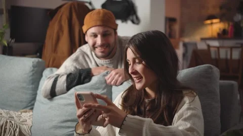 Young Hispanic couple chatting while looking at a smartphone on their home sofa, Stock Footage 346162210