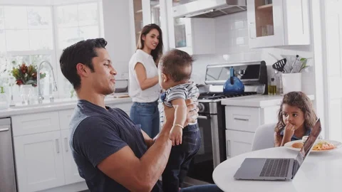 Young Hispanic family in their kitchen, mum cooking at hob, dad lifting baby in Stock Footage