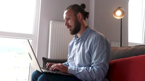 Young hispanic guy sitting on the couch, looking out the window and typing a  Stock Footage 136626074