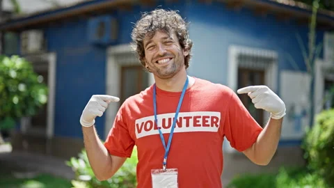 Young hispanic man activist pointing to volunteer uniform smiling at park Stock Footage 247379124