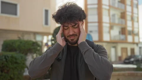 Young hispanic man with beard holding head outdoors on sunny day, standing .. 스톡 동영상 307294149