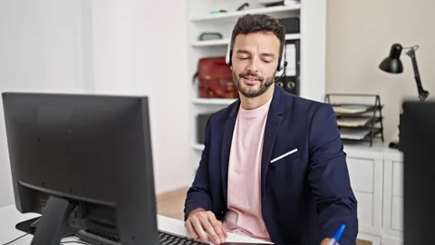 Young hispanic man call center agent having video call taking notes at office Video stock 247378819