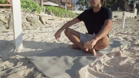 Young hispanic man with cap sits and does yoga on the beach with his yogamat Video stock 172441837