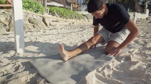 Young hispanic man with cap stretches legs on his yogamat at the beach Stock Footage 172441502