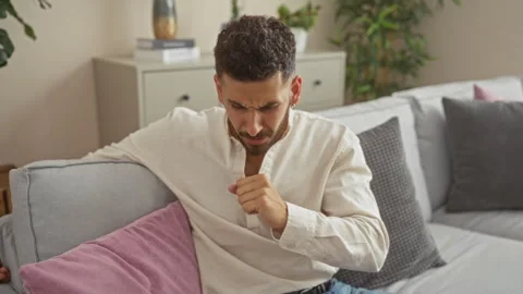 A young hispanic man coughs while sitting on a couch in a living room, port.. Stock Footage 281424367