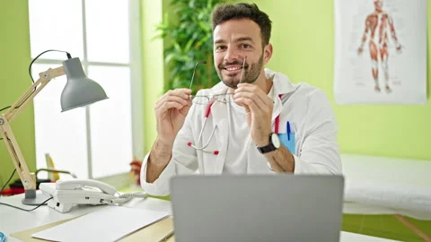 Young hispanic man doctor using laptop taking glasses off smiling at clinic Stock Footage 247378865