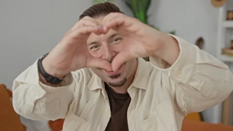 Young hispanic man forms heart shape with hands over face while seated on o.. 動画素材 339213532