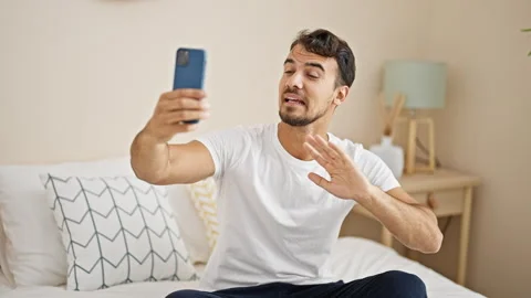 Young hispanic man having video call sitting on bed at bedroom Stock Footage 245030606