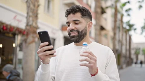 Young hispanic man holding bottle of water using smartphone at street Video stock 245047381