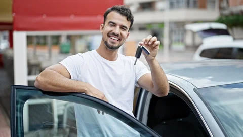 Young hispanic man holding key of new car leaning on door car at street Stock Footage 245031947
