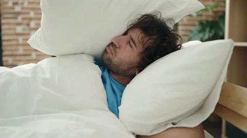 Young hispanic man lying on bed covering ears with pillow for noise at bedroo Stock Footage 232255309