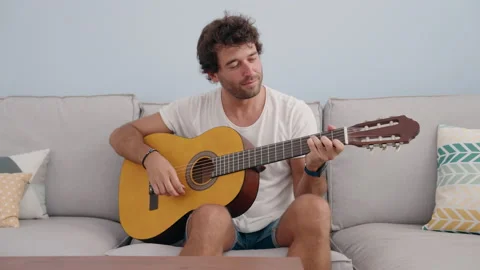 Young hispanic man playing classical guitar sitting on sofa at home Stock Footage 205020706
