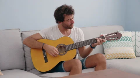 Young hispanic man playing classical guitar sitting on sofa at home Stock Footage 205524542
