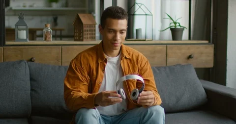 Young Hispanic man is putting wireless headset on, sitting on sofa, using gadget Stock Footage 113647837