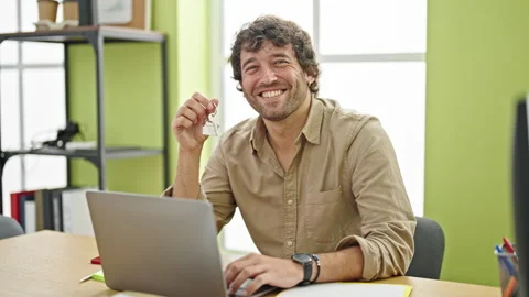 Young hispanic man real state agent holding key of new home working at office Stock Footage 247379187