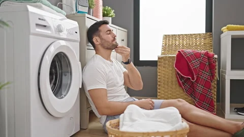 Young hispanic man sitting on floor tired waiting for washing machine at la.. Stock Footage 243489798