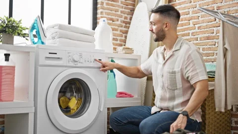 Young hispanic man sitting on floor starting washing machine looking watch .. Stock Footage 247382279